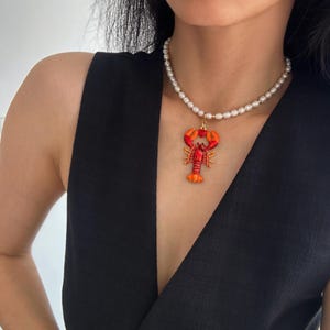 Könnte beinhalten: Eine Perlenkette mit einem Hummer-Anhänger. Der Anhänger ist rot, orange und goldfarben. Die Kette wird von einer Person getragen, die ein schwarzes Oberteil trägt.