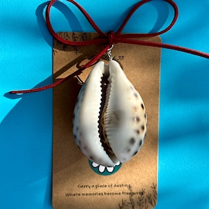 Pode incluir: Um pingente de concha de búzio com uma fiança de prata, preso a um cartão marrom com o texto "Carry a piece of destiny, Where memories become treasures." Um cordão vermelho é amarrado em um laço na parte superior. A concha é branca com manchas marrons.