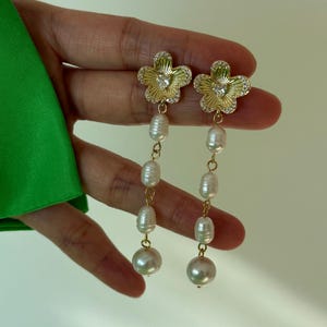 May include: Gold-tone flower earrings with dangling white pearls. The flower-shaped tops are embellished with clear crystals. The earrings have a delicate design, with a large pearl at the bottom of each earring.