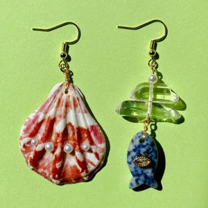 Puede incluir: Un par de pendientes únicos. Uno presenta una concha con patrones rojos y blancos y pequeñas perlas. El otro tiene un diseño claro y en capas y un dije en forma de pez azul con detalles dorados. Ambos tienen ganchos dorados.