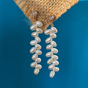 May include: Elegant gold-toned earrings featuring a cluster of white, oval-shaped pearls. Each earring has a small, round, clear gemstone at the top. The earrings hang from a gold wire and are displayed against a blue background.