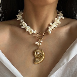May include: A statement necklace featuring a gold pendant in the shape of a nautilus shell. The necklace has a toggle clasp and is adorned with white shell fragments and small pearls. The pendant hangs from a delicate chain.