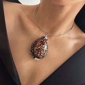 May include: A silver-toned metal choker necklace featuring a large, teardrop-shaped cowrie shell pendant. The shell has a glossy, cream-colored base with a dense pattern of dark brown, leopard-like spots covering its entire smooth surface.