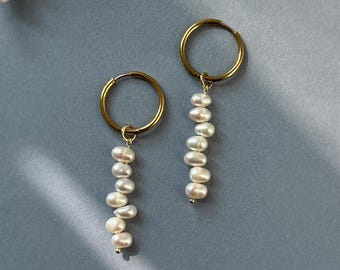 Può includere: Un paio di orecchini a cerchio color oro, ciascuno caratterizzato da un pendente verticale composto da diverse piccole perle d'acqua dolce bianche e irregolari. I cerchi sono circolari e metallici, con le perle sospese alla base.