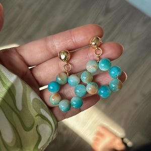 May include: A pair of dangle earrings featuring circular hoops made of polished turquoise and tan marbled stone beads. Each earring is suspended from a smooth, round gold-tone metal stud post, connected by a small gold-tone metal jump ring.
