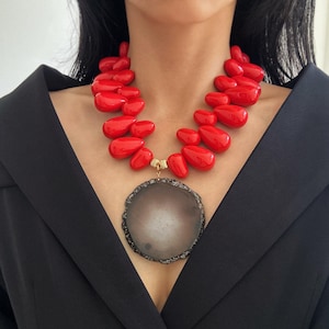 May include: A bold red statement necklace featuring large, glossy, teardrop-shaped beads. The necklace includes a large, round pendant with a gray and white center and a decorative, dark-colored border. The necklace is worn over a black garment.