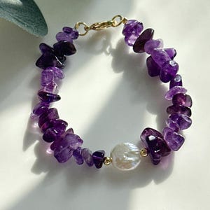 Puede incluir: Un brazalete de chips de amatista púrpura con un solo dije de perla blanca. El brazalete está hecho con piedras naturales y tiene un cierre dorado.