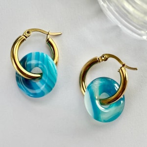 May include: A pair of gold-toned hoop earrings, each featuring a circular charm with a swirling pattern of turquoise, light blue, and white. The hoops have a hinged closure mechanism, and the charms are suspended from the bottom of each hoop.