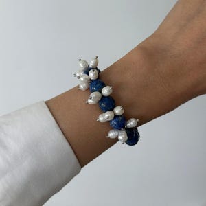 Puede incluir: Una pulsera de cuentas que alterna piedras redondas de lapislázuli azul oscuro con grupos de pequeñas perlas de agua dulce blancas e irregulares. Las cuentas están ensartadas, creando una apariencia texturizada y multicapa en la muñeca.