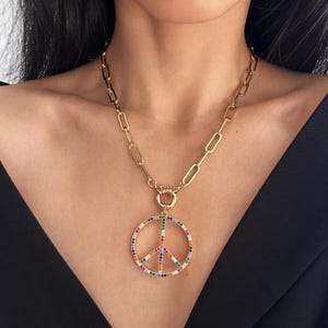 May include: Gold-coloured necklace with a peace sign pendant. The pendant is adorned with colourful gemstones. The chain has a chunky link design. The necklace is worn against a black top.