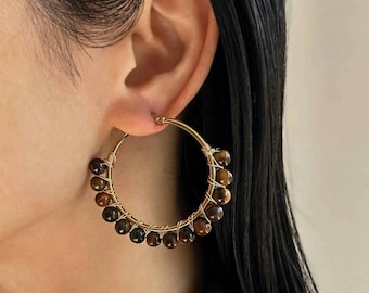 May include: A gold-toned hoop earring featuring a series of round, brown, and tan tiger's eye beads wrapped around the lower half of the wire frame. The beads are secured to the thin metal hoop with fine gold-colored wire, creating a circular design.