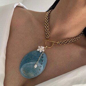 May include: A gold-toned chain necklace featuring a toggle clasp and a large, oval-shaped blue abalone shell pendant. The pendant is accented with a cluster of small white pearls and a single dangling pearl on a delicate gold-toned chain link.