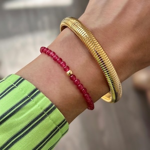 Pode incluir: Um bracelete de corrente de cobra dourada e um bracelete de contas vermelhas com um acento de contas douradas.