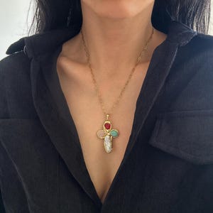 Puede incluir: Collar dorado con un colgante en forma de cruz. El colgante presenta una gran perla irregular en la parte inferior, con una piedra preciosa rosa, roja y verde en cada brazo. El collar se lleva sobre una camisa negra.