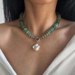 Puede incluir: Un collar de cuentas verdes con un gran colgante de perla blanca irregular. El collar tiene un cierre plateado y se lleva alrededor del cuello. Las cuentas son de color verde claro.