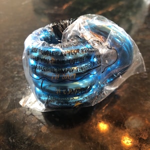 May include: A blue plastic package containing a Tangle toy. The package has the text "USE TANGLE® AS YOUR PERSONAL HELP YOU THINK "OUT OF THE BOX" TANGLE® IS SIMPLY TWISTED GOOD" and "©1981 Richard X. Zaugg" printed on it.