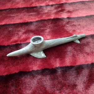 May include: A light gray ceramic pipe shaped like a submarine, featuring a cylindrical bowl on top, a tapered nose, and small side fins. The surface has a smooth, glossy finish with subtle texture details that mimic the structure of a vessel.