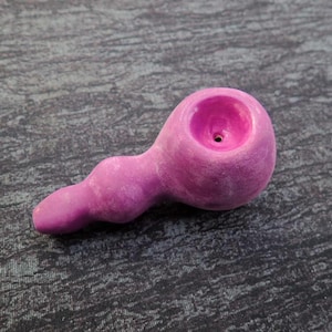 May include: A pink ceramic pipe with a rounded bowl and a curved stem. The pipe has a textured surface and a small hole in the bowl.