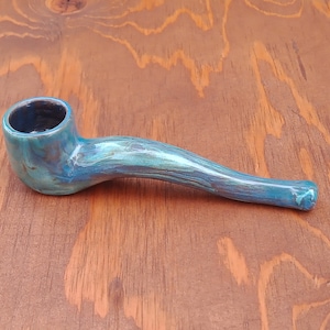 May include: A ceramic smoking pipe featuring a glossy, marbled glaze in shades of teal, turquoise, and deep blue. The pipe has a curved, elongated stem and a rounded bowl with a dark interior, showcasing a smooth, reflective finish throughout.