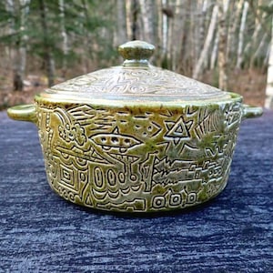 Könnte beinhalten: Ein grüner Keramik-Auflauf mit Deckel, mit geätzten Designs aus geometrischen Formen und Symbolen. Die Schüssel hat zwei Griffe und einen Knopf auf dem Deckel. Der Hintergrund ist eine verschwommene Waldszene.