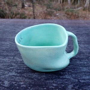 May include: A handmade, light teal ceramic mug with a unique, asymmetrical shape. The mug features a textured handle and the word "MAX" is written on the inside rim. The mug is sitting on a dark blue textured surface.