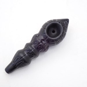 May include: A dark purple, carved pipe with intricate patterns and a bowl-shaped head. The pipe has a long, slender body with decorative ridges and a mouthpiece at the end. The design is symmetrical and detailed.