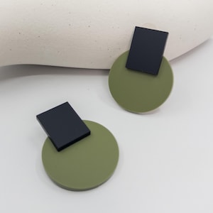 Puede incluir: Un par de pendientes modernos. Cada pendiente presenta una forma cuadrada negra unida a un círculo verde oliva. Los pendientes están colocados sobre un fondo blanco.