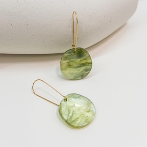 May include: A pair of earrings with a marbled green and white pattern. The earrings are circular and hang from gold-coloured ear wires. These earrings are a fashionable accessory.