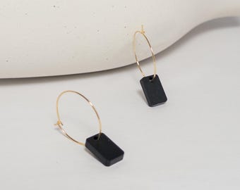 May include: A pair of gold hoop earrings with rectangular black pendants. The earrings are displayed on a white surface, with a neutral background. The earrings are a simple, modern design.