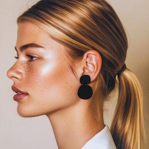 May include: A woman with long blonde hair wearing a pair of black geometric earrings. The earrings are made of two round shapes, one slightly larger than the other, connected by a small metal bar.