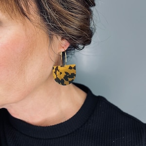 Puede incluir: Un par de pendientes de aro dorados con un colgante en forma de semicírculo. El colgante es marrón y negro con un patrón moteado.