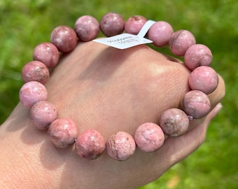 May include: A pink rhodonite gemstone bracelet. The bracelet features round, polished beads in varying shades of pink and brown. The bracelet is held in a hand against a blurred green background.