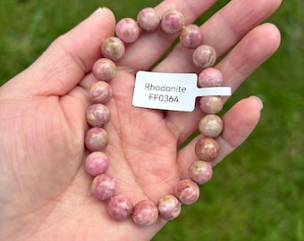 May include: A pink rhodonite gemstone bracelet. The bracelet is composed of round, polished beads in shades of pink and tan. A white tag with the text "Rhodonite FF0364" is attached to the bracelet.