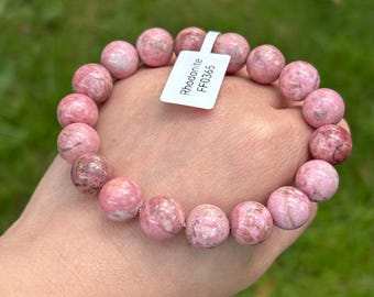 May include: A pink rhodonite bracelet with round beads. The bracelet is held in a hand against a green background. A white tag with the text "Rhodonite FF0365" is attached to the bracelet.