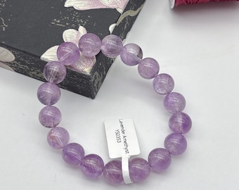 Puede incluir: Un brazalete de cuentas de amatista lavanda. El brazalete presenta cuentas redondas y translúcidas en diferentes tonos de púrpura. Se adjunta una etiqueta blanca con el texto "Lavender Amethyst YS0353".
