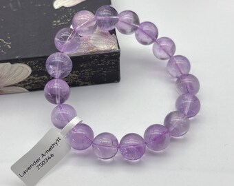 Puede incluir: Un brazalete de cuentas de amatista lavanda. El brazalete presenta grandes cuentas redondas y translúcidas en varios tonos de púrpura. Una etiqueta blanca dice "Lavender Amethyst ZS0346". El brazalete se exhibe sobre una superficie blanca.