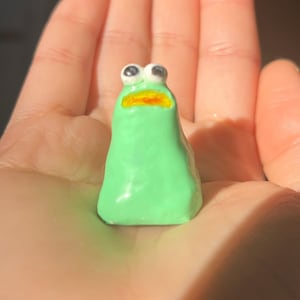 May include: A small green ceramic figurine with large black eyes and a yellow mouth. The figurine is shaped like a slug.