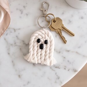May include: A ghost-shaped keychain crafted from off-white twisted cotton rope. The ghost features two small black oval eyes and one larger black oval mouth. It is attached to a silver-toned metal lobster clasp and a split ring holding two brass keys.