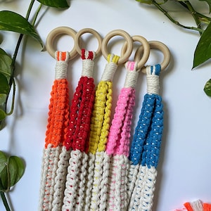 Puede incluir: Cinco maceteros de macramé con anillos de madera. Los maceteros están hechos con cordón de color crema natural y tienen detalles coloridos en naranja, rojo, amarillo, rosa y azul.
