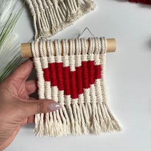 Può includere: Un piccolo arazzo da parete con un motivo a forma di cuore rosso intrecciato su uno sfondo di macramè bianco. L'arazzo è fissato a un bastone di legno.