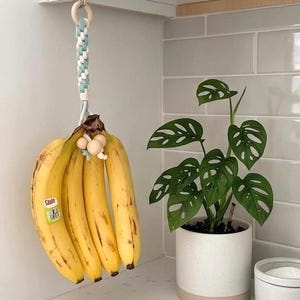 Peut inclure: Un porte-bananes en macramé avec une corde tressée bleu clair et blanche, un anneau en bois sur le dessus et des perles en bois à la base. Le support est suspendu à un crochet et maintient un régime de bananes jaunes avec une étiquette verte.