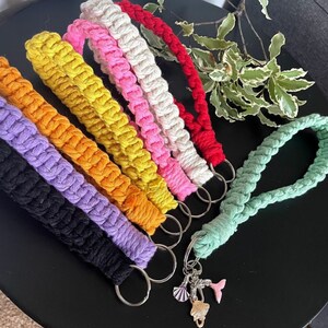 Puede incluir: Un conjunto de llaveros de macramé de colores en una variedad de colores que incluyen negro, blanco, amarillo, naranja, morado, rosa y verde. Los llaveros están unidos a llaveros de plata con un dije unido a uno de los llaveros.