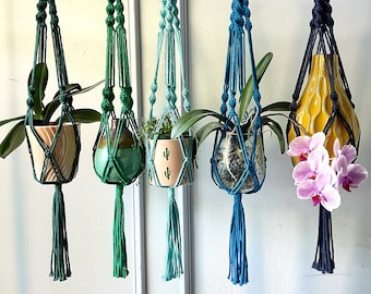 May include: Five macramé plant hangers in shades of green, blue, and yellow. Each hanger suspends a different plant or vase, including orchids and a cactus. The hangers are suspended from wooden rings, with decorative tassels at the bottom.