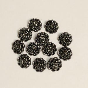 May include: A collection of eleven small, metallic, gunmetal-coloured buttons shaped like blooming roses. Each button features a detailed, swirling petal design with a raised centre, creating a textured, circular appearance for garment fastening.