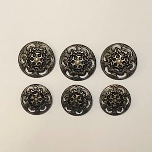 May include: A set of six round metal buttons featuring an intricate, raised filigree pattern with a dark, antiqued bronze finish. Each button displays a symmetrical, openwork design with a central star-like motif surrounded by scrolling details.