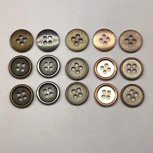 May include: A collection of fifteen round, four-hole buttons arranged in three rows of five. The buttons feature various metallic finishes, including antique brass, dark gunmetal, copper, and silver tones, with some having raised rims.