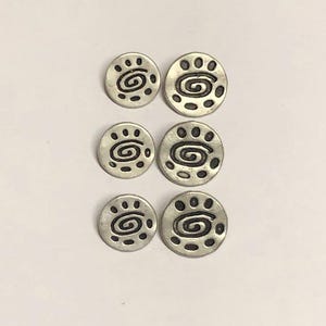 May include: A set of six circular silver-toned metal buttons arranged in two columns. Each button features a central spiral design surrounded by a series of small, dark, recessed dots along the outer edge, creating a consistent pattern across all pieces.