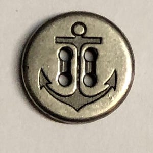May include: A round, metallic-toned button featuring a dark, engraved anchor design centered on its surface. The button includes four holes arranged vertically in the center of the anchor, providing a functional attachment point for sewing projects.