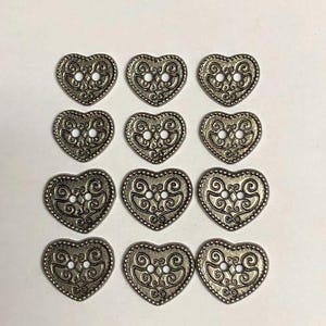 May include: A set of twelve silver-toned metal buttons, each shaped like a heart with a raised beaded border. The surface of each button features an intricate embossed scrollwork pattern and two central holes for attachment to fabric or garments.