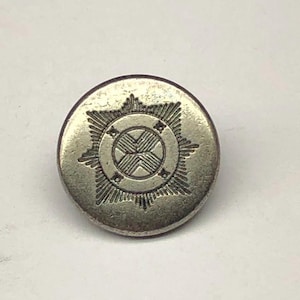 May include: A circular silver-toned metal button featuring an embossed starburst design. The center of the button displays a circular emblem with a cross-hatched pattern, surrounded by a radiating star shape with pointed rays extending outward.